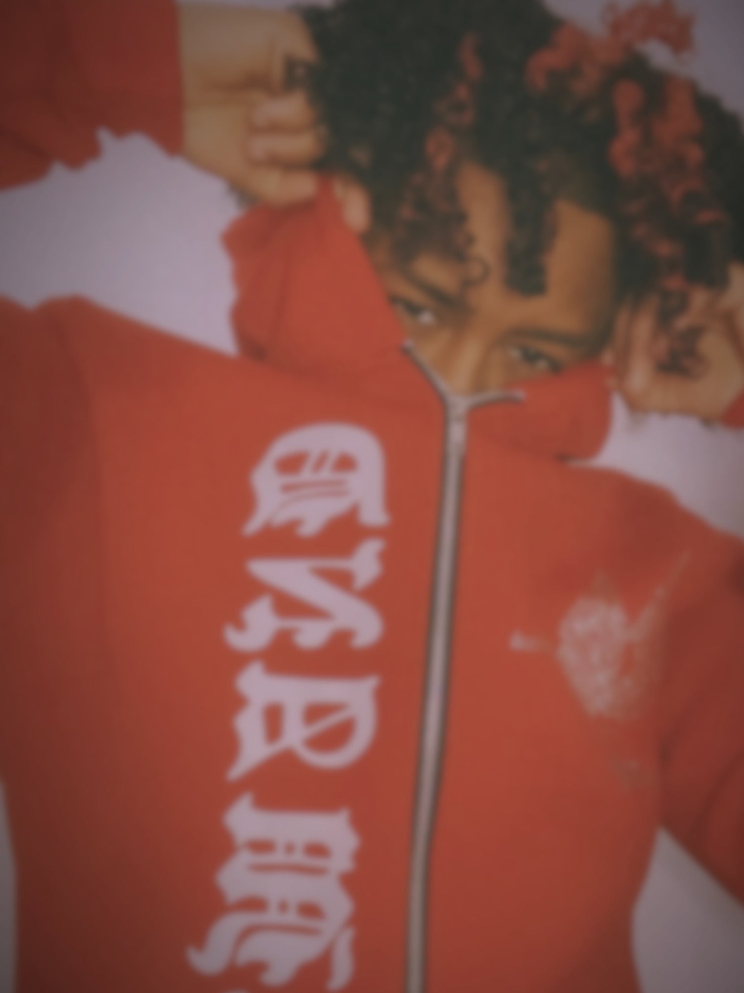 A person wearing a bright red zip-up hoodie with large white gothic-style lettering down the front, posed against a white background.